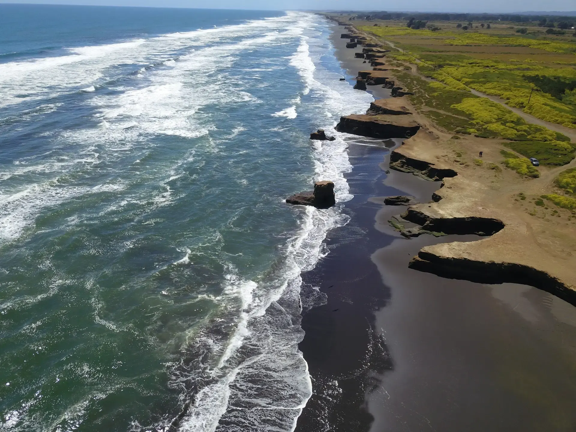 Vista aérea de la costa: playa, oleaje y acantilados junto a terrenos de campo